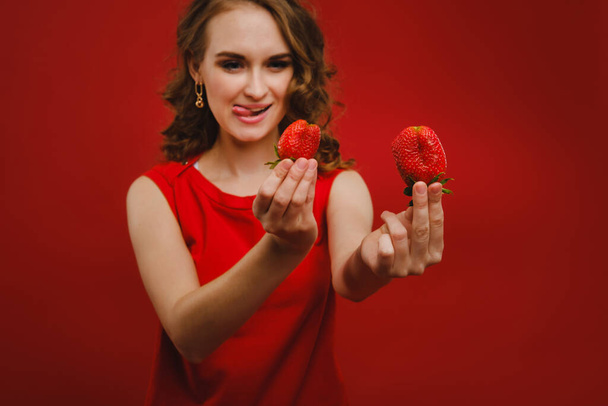 Piękna dziewczyna w czerwonej sukience na czerwonym tle trzyma truskawkę w dłoniach i uśmiecha się - Zdjęcie, obraz