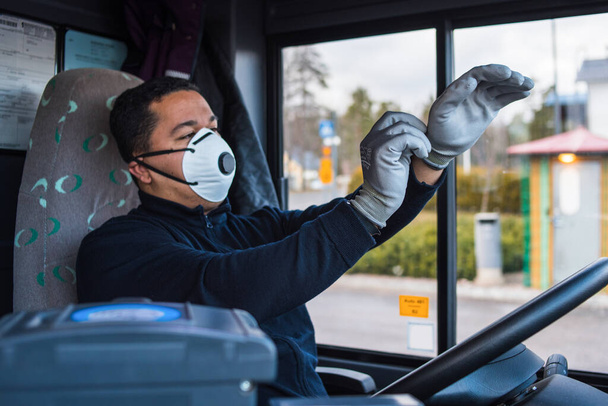 Der junge hispanische Busfahrer mit Maske zieht sich im Bus Schutzhandschuhe an die Hand, um sich vor der Coronavirus-Epidemie zu schützen. Covid 19. Schutz vor Coronaviren. Quarantäne 2020. zu Hause bleiben. - Foto, Bild