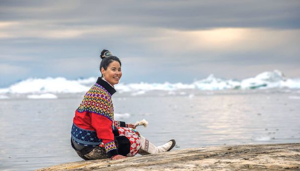 Ilulissat, Greenland - July 11, 2018: Young inuit woman in traditional clothing posing for photos on the shore of the ocean, nearby by small Greenlandish village. - Фото, изображение