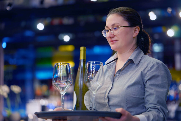 Číšník obsluhuje stůl. Servírka nosí na podnose v hotelové restauraci v baru skleničky na víno a lahvové víno. Koncept služby. - Fotografie, Obrázek