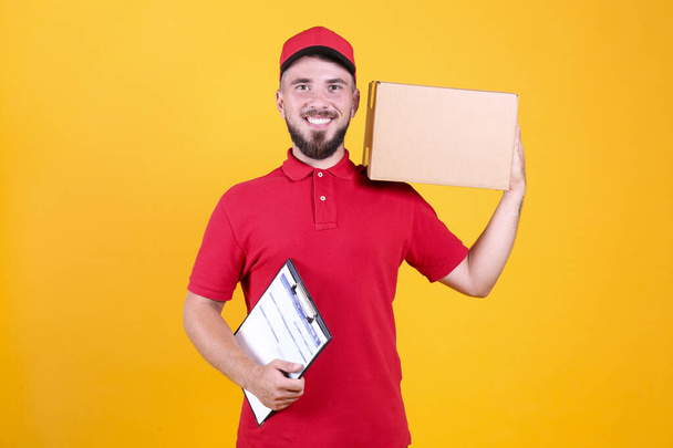 Young handsome delivery guy wearing red uniform and cap holding the blank cardboard box over isolated white background. Portrait of friendly bearded man carrying parcel. Copy space for text. - Fotografie, Obrázek