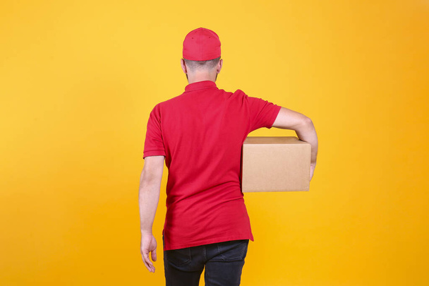 Back view of young delivery guy wearing red uniform and cap holding the blank cardboard box over isolated yellow background. Back view of bearded man carrying parcel. Copy space for text. - Fotografie, Obrázek