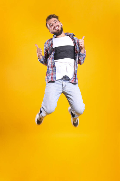 Young handsome man with facial hair jumping over yellow wall with a lot of copy space for text. Portrait of confident bearded male, wearing fashionable casual outfit. Isolated, background. - Foto, imagen