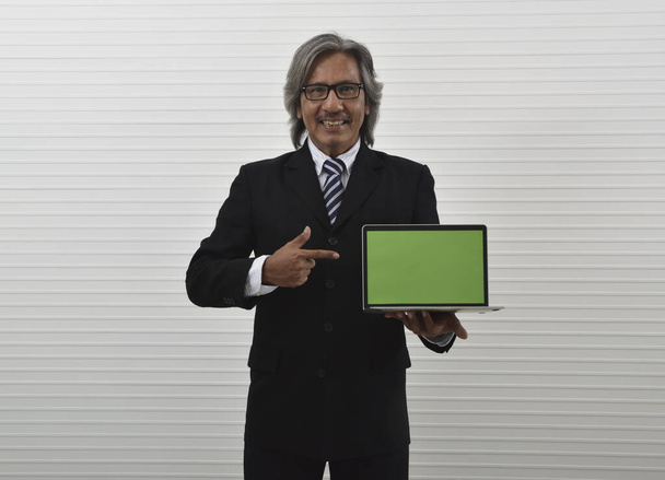 Happy elderly asian businessman in black suit and eyeglasses holding laptop computer and pointing finger to screen standing over white wall background, Business internet online concept - Photo, image