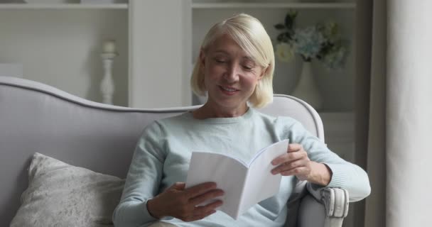 Happy mature elderly woman resting on couch, reading book. - Video, Çekim