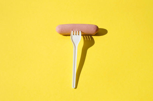 Salchicha y tenedor de plástico blanco sobre fondo amarillo brillante con luz dura
 - Foto, imagen