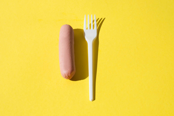 Salchicha y tenedor de plástico blanco sobre fondo amarillo brillante con luz dura
 - Foto, Imagen