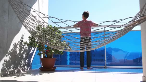 Kid on balcony looks around, lies down in the hammock and swings. Stay home, social distance, self-isolation during Coronavirus quarantine - Footage, Video
