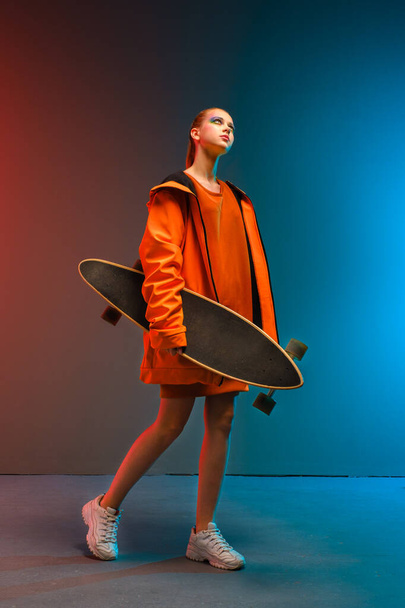 A young girl with bright makeup holds a longboard in her hands on a fashionable neon color background. Portrait of a girl skater - Zdjęcie, obraz