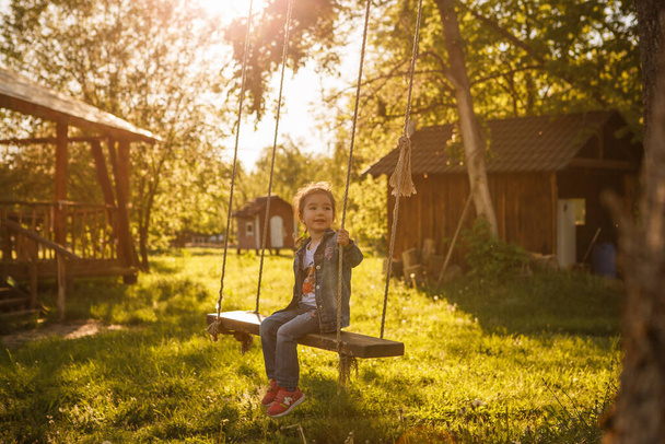 A little girl in a denim suit swings on a suspended swing to a tree. A girl in blue jeans on a swing with long ropes - Zdjęcie, obraz