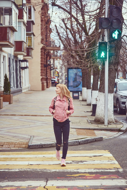 Ένα κορίτσι με ροζ μπουφάν στην πόλη διασχίζει τον δρόμο διασχίζοντας πεζούς. Το φανάρι είναι πράσινο.. - Φωτογραφία, εικόνα