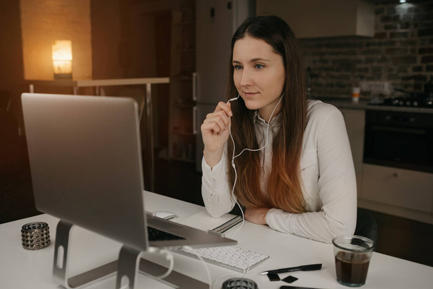 Fernbedienung. Eine kaukasische Frau mit Kopfhörern, die mit ihrem Laptop ferngesteuert online arbeitet. Ein Mädchen im weißen Hemd diskutiert mit ihren Kollegen per Videoanruf an ihrem gemütlichen Arbeitsplatz zu Hause über Geschäfte. - Foto, Bild