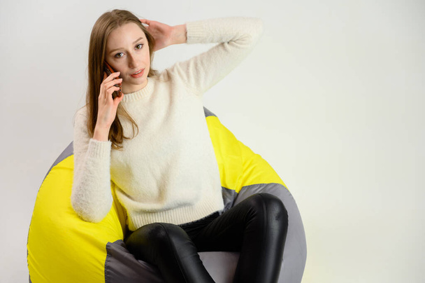Studio portrait of a happy caucasian girl in a white sweater sitting on a white background talking with a phone in her hands - Foto, Imagen