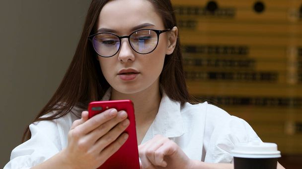 Młoda dziewczyna w okularach używa smartfona, zbliżenie. Student trzyma w rękach czerwony telefon. - Zdjęcie, obraz