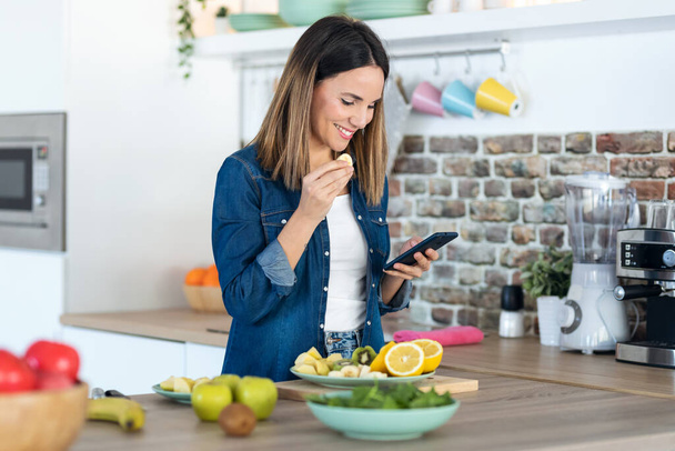 Tiro de mulher bonita usando seu telefone celular enquanto come uma fatia de banana na cozinha em casa
. - Foto, Imagem