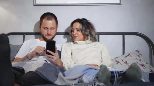Ευτυχισμένο ζευγάρι αγκαλίτσες στο κρεβάτι με smartphone στο σπίτι στο υπνοδωμάτιο. Νεαρό ζευγάρι που χρησιμοποιεί έξυπνα τηλέφωνα στο κρεβάτι - Πλάνα, βίντεο