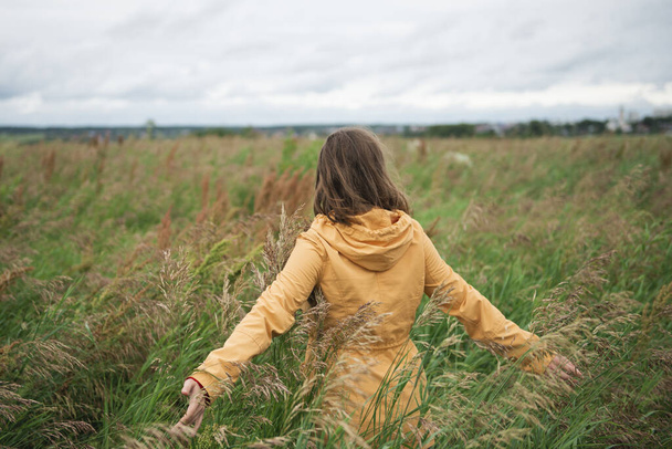 Young woman in yellow jacket with opened arms standing in field against cloudy sky looking at horizon - Фото, изображение