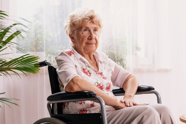 Elderly ailing woman dressed in a blouse with flower pattern is sitting in a wheelchair in her home - Photo, Image