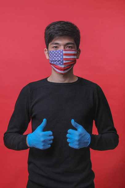Un joven con una máscara médica con la bandera de Estados Unidos, guantes médicos azules, sobre un fondo rojo, muestra con sus dos manos "todo está bien" en la lente. máscara médica. guantes médicos
 - Foto, imagen