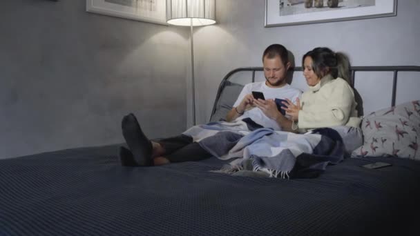 Высокая дуга снимка молодого кавказского мужа и жены в пижамах, сидящих молча в постели ночью, опирающихся на спинку кровати, держащих смартфоны и выглядящих полностью погруженными - Кадры, видео