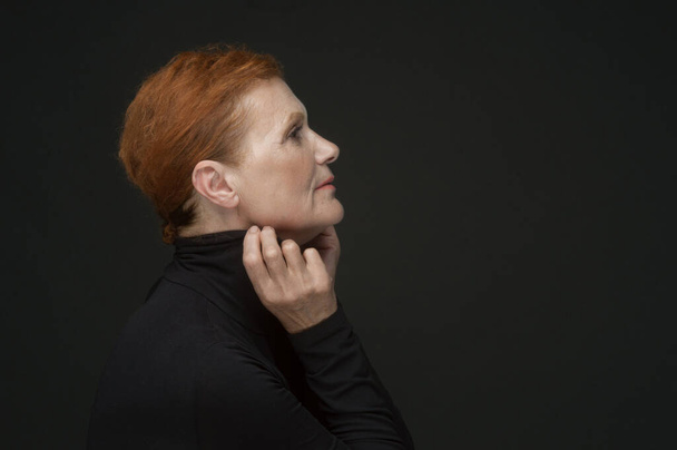 Profile portrait of a red-haired elderly lady in a black sweater on a dark gray background - Fotó, kép