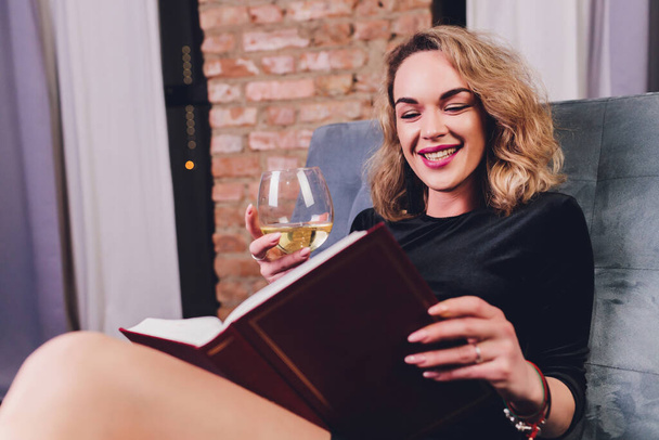 Csinos fiatal vörös hajú nő olvas egy könyvet fekvő fekete bőr kanapén egy pohár vagy vörösbor. - Fotó, kép