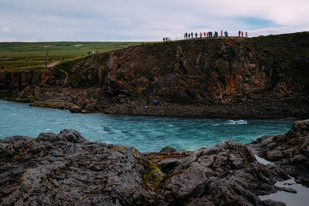 Blick auf den Fluss und Touristenmassen in der Nähe eines Wasserfalls in Island in der Nähe des Myvatn-Sees im Sommer an einem bewölkten Tag - Foto, Bild
