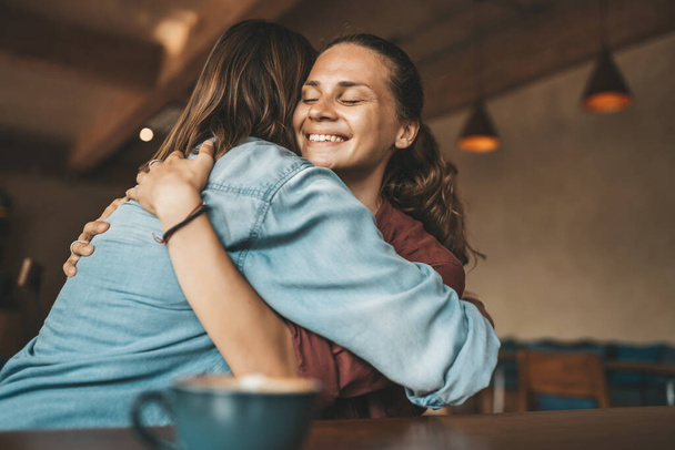 Ευτυχισμένη νεαρή γυναίκα αγκαλιάζει τον καλύτερό της φίλο, κάθεται σε ένα καφέ, έχοντας ευχάριστη συζήτηση. - Φωτογραφία, εικόνα