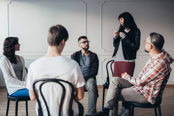 Sad teenager stands in front of a group of people of different ages during a therapy group meeting and talks about his problems - Photo, Image