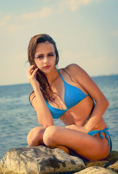young brunette girl in a bright blue bathing suit sits on a large stone against the sea. Copy space - Φωτογραφία, εικόνα