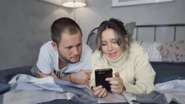 mies ja nainen makaavat sängyssä ennen nukkumaanmenoa, käyttäen älypuhelimia, näyttämällä kuvia - Materiaali, video