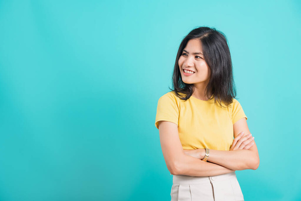 Portrait Asian beautiful happy young woman smiling wear yellow t-shirt standing cross arm with looking side away at copy space, shoot photo in a studio on a blue background - Foto, afbeelding