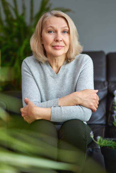 Lovely middle-aged blond woman sitting on a sofa at home looking at the camera. - Fotografie, Obrázek