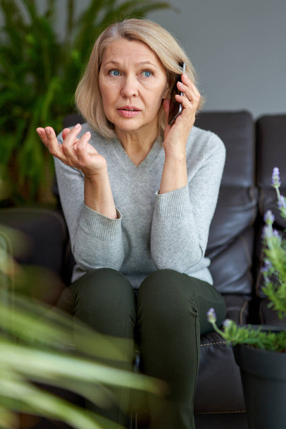 Mujer seria asistiendo a una llamada telefónica sentada en un sofá en la sala de estar en casa
. - Foto, imagen