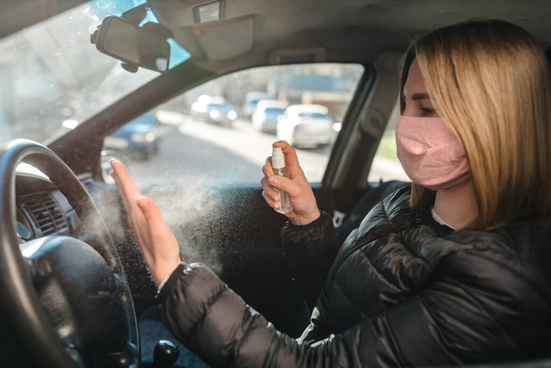 Spraying antibacterial sanitizer spray on hand in car, infection control concept. Sanitizer to prevent Coronavirus, COVID-19, flu. Spray bottle. Woman wearing in medical protective mask driving a car. - Foto, imagen