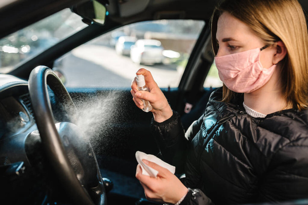 Spray antybakteryjny spray dezynfekujący na kierownicę, samochód dezynfekujący, koncepcja kontroli infekcji. Zapobiegać koronawirusowi, COVID-19, grypie. Kobieta nosząca medyczną maskę ochronną prowadzącą samochód. - Zdjęcie, obraz