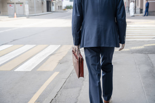 Einsamer Geschäftsmann im blauen Anzug mit Aktentasche und weißen Gummihandschuhen auf dem Fußgängerüberweg an der Stadtstraße. Gesundheitsschutz vor Viren, Keimen und Bakterien. Coronavirus - Foto, Bild