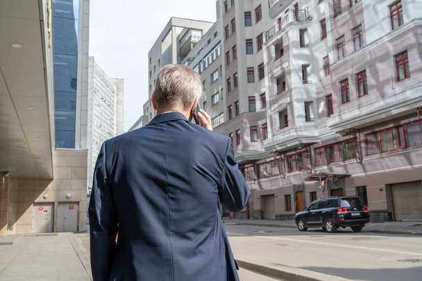 Solo hombre de negocios de mediana edad de pelo gris en un traje azul llamando por teléfono de pie en la calle vacía de la ciudad
. - Foto, imagen