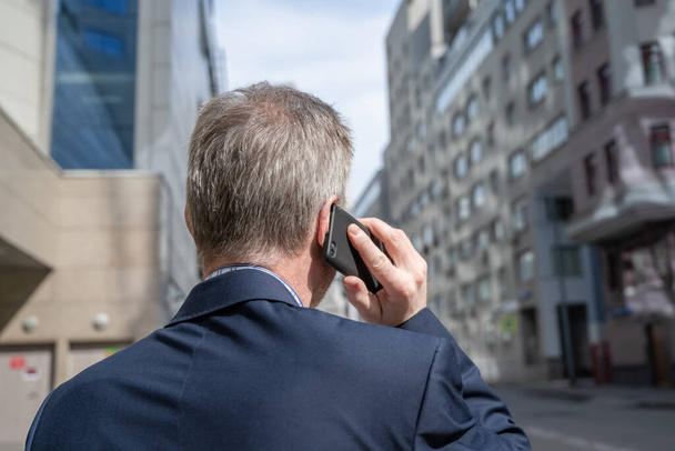 Alone middle-aged gray-haired businessman in a blue suit calling by phone standing on the empty city street. - Photo, image