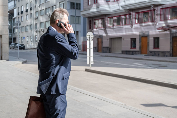 Один середнього віку сіро-волохатий бізнесмен в синьому костюмі дзвонить по телефону, стоячи на порожній міській вулиці
. - Фото, зображення