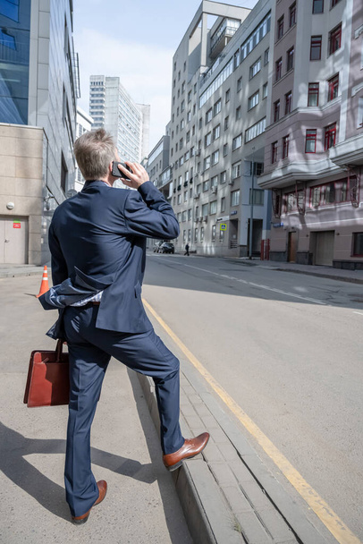 Solo hombre de negocios de mediana edad de pelo gris en un traje azul con maletín llamando por teléfono camina en el paso de peatones en la calle de la ciudad
. - Foto, imagen