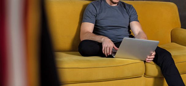 Casual handsome young man relaxing at home sitting on the couch and works on a laptop. Man in blank shirt for you mockup - Foto, afbeelding