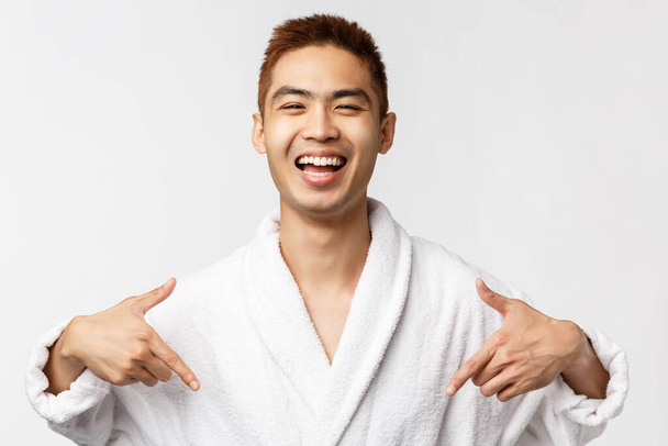 Krása, lázně a volný čas koncept. Portrét spokojený, šťastný asijský muž v županu ukazuje na sebe a široce se usmívá, mluví vlastní úspěch, osobní úspěch, bílé pozadí - Fotografie, Obrázek