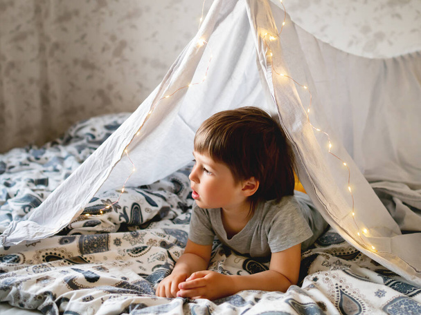 Little boy plays with his teddy bear. Toddler lying in tent made of linen sheet on bed. Cozy evening with favorite toy in DIY interior. - Photo, Image