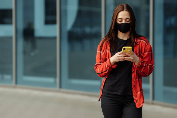 Junge schöne Frau trägt schwarze Gesichtsmaske Schutz gegen epidemische Coronavirus covid-19. Sieht auf dem Bildschirm des Telefons aus. Europäische Mädchen medizinische Schutzmaske orange Business-Anzug - Foto, Bild
