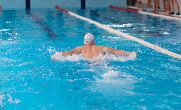 Zwembad atleet training binnen voor professionele competitie. tiener zwemmer dragen witte badmuts uitvoeren van de vlinder slag - Foto, afbeelding