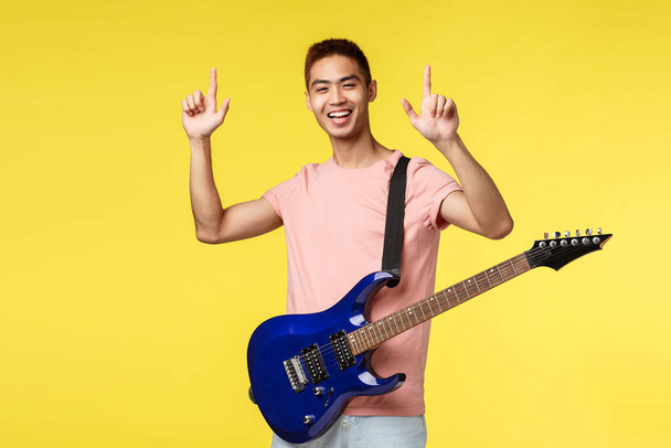 Životní styl, volný čas a mládež koncept. Šťastný nadšený hezký mladý asijský muž ukazuje prsty nahoru, vystupuje na jevišti, drží elektrickou kytaru a usmívá se, žluté pozadí - Fotografie, Obrázek