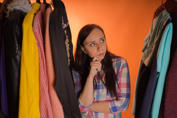Young woman grimacing standing between clothes in wardrobe. Adult female choosing things in store. - Photo, Image