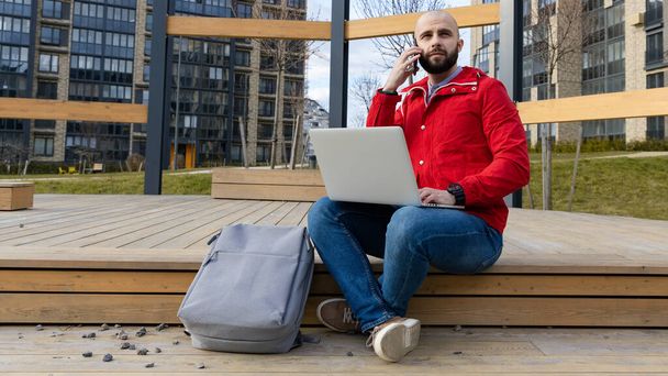 Ένας βάναυσος τύπος με γενειάδα σε καθημερινά ρούχα δουλεύει σε έναν υπολογιστή και μιλάει στο τηλέφωνο στο δρόμο. Έννοια της εργασίας εκτός σπιτιού - Φωτογραφία, εικόνα