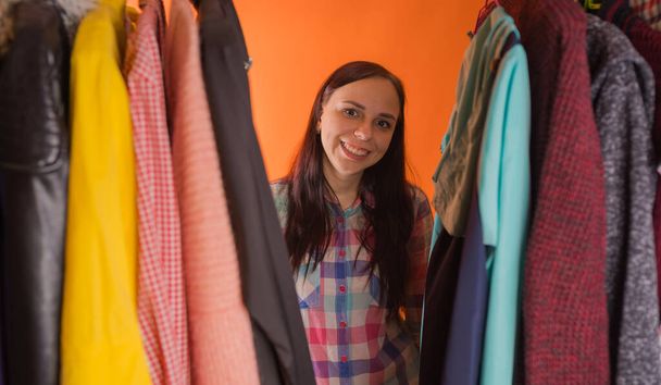 Mujer joven haciendo muecas de pie entre la ropa en el armario. Mujer adulta eligiendo cosas en la tienda
. - Foto, imagen